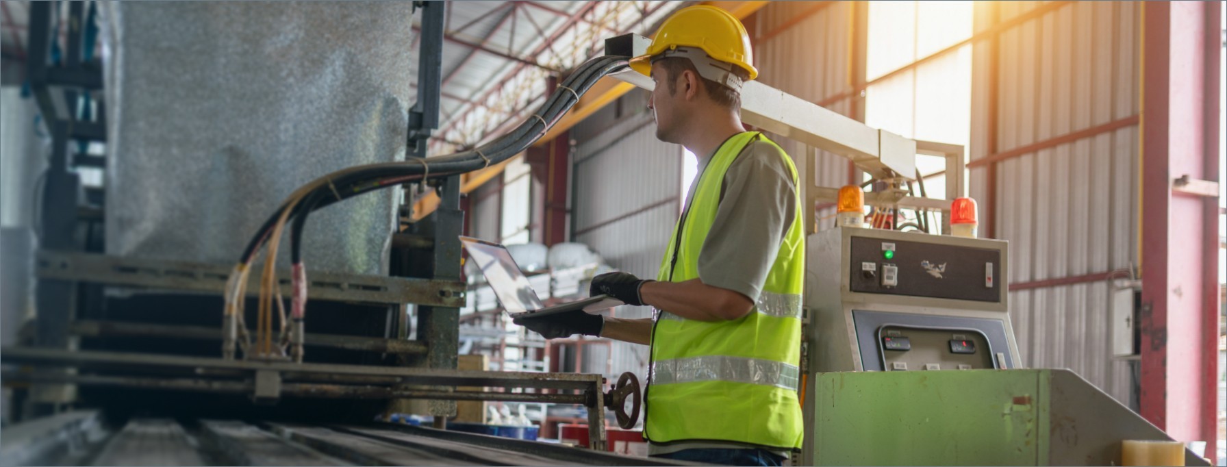 Operaio in un impianto industriale con elmetto giallo e gilet ad alta visibilità, che utilizza un laptop davanti a macchinari di produzione.